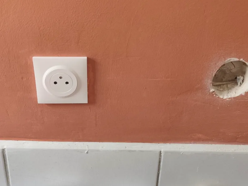 Prise électrique neuve posée sur un mur peint, à côté d'une boîte d'encastrement en préparation